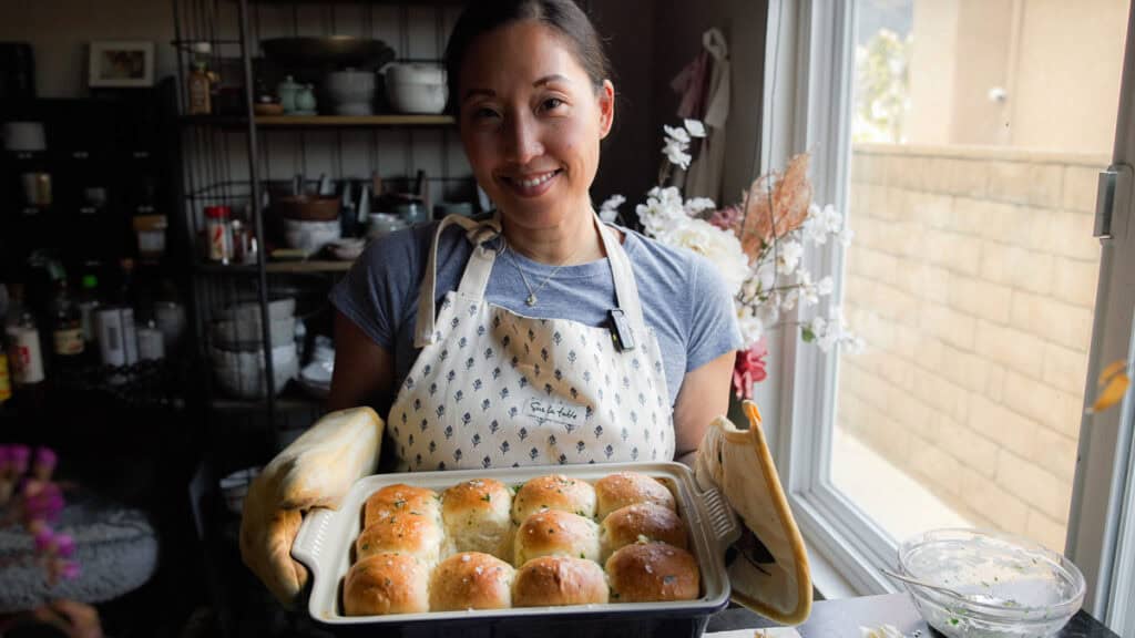 A person wearing an apron holds a tray of freshly baked vegan dinner rolls. They are standing in a cozy kitchen by a window, with shelves filled with jars and kitchen items in the background.